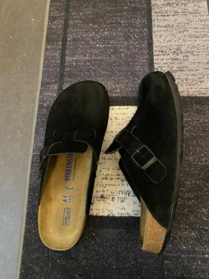 Svarta Birkenstock tofflor i mocka - Säljer ett par svarta Birkenstock tofflor i mocka med justerbart spänne och klassisk korksula. Modellen har öppen häl och mjuk innersula för extra komfort. Perfekta för chill dagar hemma eller när du vill ha något bekvämt på fötterna.