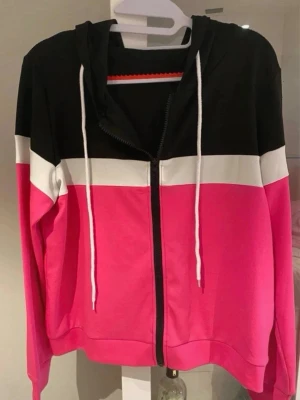 Färgglad hoodie med dragkedja - Säljer en  tunnare hoodie med dragkedja i färgblock: svart upptill, vit rand i mitten och starkt rosa nedtill och på ärmarna. Tröjan har huva med vita snören och är tillverkad i mjukt sweatshirtmaterial. Perfekt för dig som gillar färg och vill sticka ut. Storlek L men är mer som M.