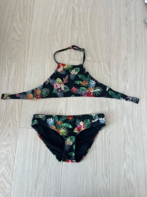 Svart blommig bikini  - Snygg svart bikini med tropiskt blommönster i grönt, rött, gult och lila. Biliniöverdelen har ett knäppe som man knäpper bak på ryggen. Bikinitrosorna har samma färgglada blommiga print. Perfekt för sommaren och strandhäng!