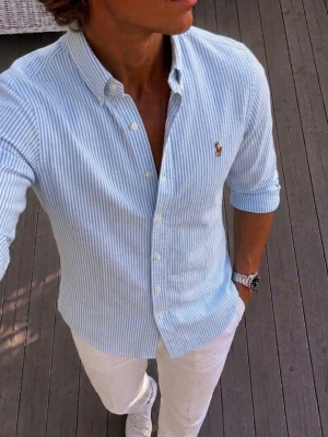 Blårandig skjorta från Ralph Lauren - Säljer en klassisk blårandig skjorta från Ralph Lauren med button-down krage och broderad logga på bröstet. Skjortan har långa ärmar, vita knappar och är tillverkad i bomull. Perfekt för en clean och stilren look.