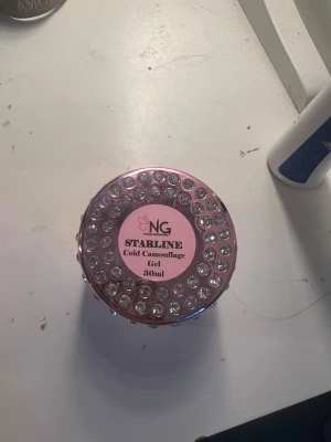 NG Starline Cold Camouflage Gel 30ml - Redan nerprutad !!!Säljer en NG Starline Cold Camouflage Gel för naglar, 30ml. Burken är dekorerad med glittrande stenar och har en rosa etikett. Gelen har en naturligt rosa ton och används för att bygga och forma naglar. Perfekt för dig som vill ha snygga och hållbara naglar.