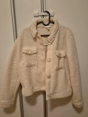 Krämvit teddyjacka med fickor strl M - Mysig krämvit teddyjacka med två bröstfickor och stora knappar. Jackan passar till mycket och är bra runt vår och höst 