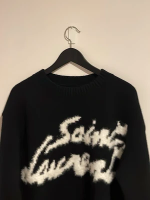 Svart stickad Saint Laurent-tröja - Säljer en svart stickad tröja från Saint Laurent med stor, fluffig vit logga framtill. Tröjan har lång ärm, ribbade muddar och en avslappnad passform. Perfekt för dig som vill ha en statement piece i garderoben.