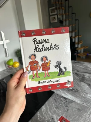 Barna Hedenhös av Bertil Almqvist - Barna Hedenhös är en illustrerad barnbok med hårda pärmar och färgglada teckningar på framsidan. Boken har en vit och röd omslagsdesign med motiv av två barn och en hund i stenåldersmiljö. Perfekt för dig som gillar klassiska svenska berättelser.