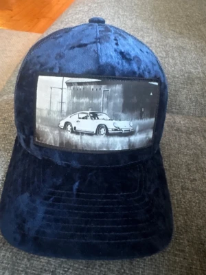 Calza Pennnello caps  - Säljer en mörkblå truckerkeps med mesh baktill och justerbar snapback. Framtill finns en stor patch med svartvitt motiv av en klassisk sportbil framför en industribyggnad. Kepsen har en cool, retro känsla och passar perfekt för dig som gillar bilar och streetwear.