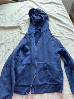 Mörkblå hoodie med dragkedja - En mörkblå hoodie med dragkedja och stor huva. Tröjan har en klassisk känguruficka framtill och ett litet rött broderat märke på bröstet. Tillverkad i mjukt sweatshirtmaterial, perfekt för chill dagar. Långa ärmar och ribbade muddar.