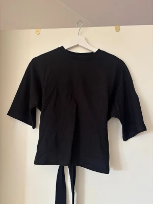 Svart topp med knytband bak - Svart topp i bomull med halvlånga ärmar och rund hals. Toppen har ett snyggt knytband i ryggen som ger en cool detalj och en croppad look baktill. Perfekt till jeans eller kjol för en trendig stil.