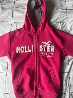 Rosa Hollister hoodie med dragkedja - Snygg rosa hoodie från Hollister med dragkedja framtill och broderad logga samt texten '1922' på bröstet. Hoodien har två fickor fram och ribbade muddar vid ärmslut och nederkant. Insidan av huvan är fodrad i grått tyg för extra komfort.