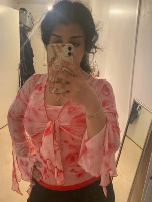 Rosa y2k blus med knyt framtill - Säljer en söt blus i tunn mesh med rosa och röda blommor. Blusen har långärmade, utsvängda ärmar och en stor knytning framtill. Materialet är genomskinligt och lätt, perfekt för lager på lager. Supersnygg till både jeans och kjol!