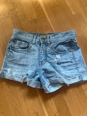 Ljusblåa denimshorts med slitningar - Snygga ljusblåa jeansshorts med hög midja och råa kanter. Shortsen har slitna detaljer både fram och bak samt klassiska fem fickor. Dom är aldrig använda, men tyvärr sitter inte prislappen kvar. Dom är i storlek S.