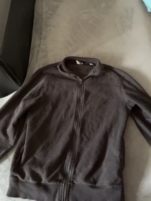 Svart zip hoodie från jack and jones - En enkel och stilren mörkbrun hoodie med dragkedja framtill och två fickor. Modellen har ribbade muddar vid ärmslut och nederkant samt en klassisk krage utan huva. Perfekt för en avslappnad look och lätt att matcha med andra plagg.