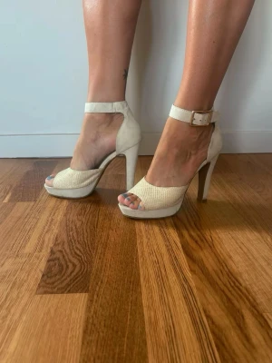 Beige höga platåpumps med spänne - Snygga beige pumps med hög klack och platåsula. Skorna har öppen tå, bred rem över foten och justerbart spänne runt ankeln. Materialet ser ut att vara mockaimitation med textur på framsidan. Perfekta för dig som vill ha extra höjd och stil.