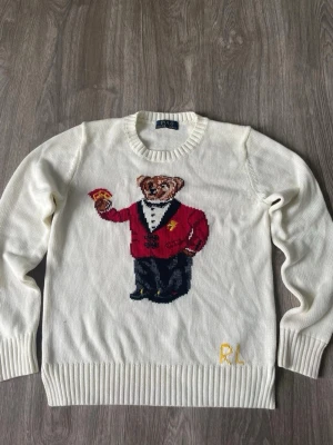 Vit stickad tröja med björnmotiv - Vit stickad tröja med rund hals och ribbade muddar. På framsidan finns ett stort motiv av en björn i röd kavaj och svart byxa, samt broderade initialer 'RL' i gult nedtill. Tröjan har en klassisk och lekfull design, perfekt för dig som gillar statement-plagg.