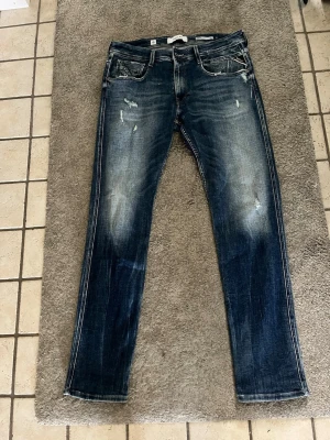 Blå jeans från Replay - Säljer ett par blå jeans från Replay med snyggt slitna detaljer och tvättad look. Modellen har raka ben, klassiska fem fickor och tydliga kontrastsömmar. Perfekta för dig som gillar en avslappnad och trendig stil. Helt nya använda en gång då dom är för stora, nypris 1700 säljer för 649kr. Storlek: W34 L32