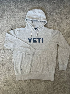Grå hoodie från YETI - Snygg grå hoodie från amerikanske Premiummärket YETI. Tröjan har huva med vita dragsnören och en klassisk magficka framtill. Tillverkad i mjukt och stabilt sweatshirtmaterial och har ribbade muddar vid ärmar och nederkant. Perfekt för en avslappnad men snygg stil. Tröjan är köpt utomlands och finns inte tillgänglig i Sverige. Premiumtillverkad och unik. Säljes till högsta budare så det är först till kvarn!