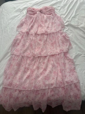 Ljusrosa axelbandslös långklänning - Romantisk axelbandslös långklänning i ljusrosa med volanger och blommigt mönster. Klänningen har flera lager i ett tunt, svepande material och en twistad detalj vid bysten. Perfekt för sommarens alla tillfällen.