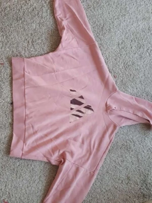 Adidas rosa hoodie - Vet inte var snörena till luvan är.  En rosa hoodie med långa ärmar och en rund halsringning. Tröjan har en lös passform och en kortare modell. På framsidan finns en diskret mönstrad detalj i form av en triangel. Ärmsluten och nederkanten är ribbade.