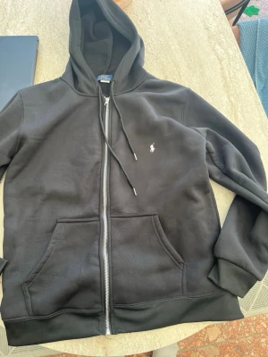 Svart hoodie från Polo Ralph Lauren - Snygg svart hoodie från Polo Ralph Lauren med dragkedja framtill, två fickor och klassisk huva med dragsko. Liten vit logga broderad på bröstet. Tillverkad i mjukt och bekvämt material, perfekt för en avslappnad stil.