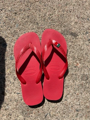 Röda flip-flops från Havaianas - Snygga röda flip-flops från Havaianas med klassisk design och bekväm sula i gummi. Remmarna har en liten detalj med flagga i rött, grönt och vitt. Perfekta för sommaren och stranden. Den portugisiska flaggan är avtagbar. Går även att köpa en annan flagga på olika butiker. Köpta i Saint Tropez på havaianas butiken