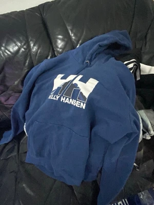 Blå Helly Hansen hoodie med tryck - Säljer en blå hoodie från Helly Hansen med stor vit logga och tryck på bröstet. Klassisk huva med dragsko och magficka framtill. Perfekt för chill dagar eller när du vill ha en sportig look.