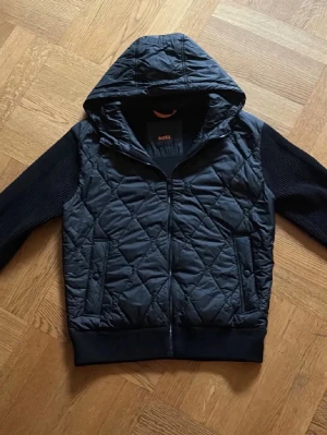 Svart cardigan från Boss  - Snygg svart hoodiejacka från BOSS med quiltat mönster på fram- och baksida samt stickade ärmar. Jackan har dragkedja framtill och en stor huva. Perfekt för dig som vill ha en stilren och bekväm jacka med streetkänsla. Använd ett fåtal gånger och sitter perfekt till storlek S