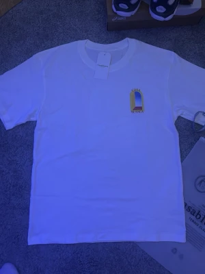 Vit t-shirt från Casablanca - Vit t-shirt från Casablanca med tryck av ett gult fönster och blå himmel på bröstet. Klassisk passform och rund hals. Tillverkad i mjuk bomull och har en clean look med diskret logga. Perfekt för dig som gillar stilrena plagg med en twist.
