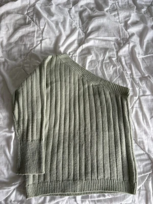 Ljusgrön ribbad one shoulder stickad tröja - Trendig ljusgrön stickad tröja med ribbad struktur och one shoulder-design. Tröjan har långa ärmar och bred mudd vid ärmslut och nederkant. Perfekt för dig som vill ha en unik och stilren look.