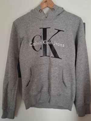 Grå hoodie från Calvin Klein Jeans - Snygg grå hoodie med stor CK-logga och texten Calvin Klein Jeans på bröstet. Klassisk känguruficka framtill, ribbade muddar och huva med metallöljetter. Perfekt för en avslappnad och trendig look. Mjuk bomullsblandning som känns skön mot huden.