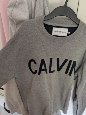 Grå sweatshirt från Calvin Klein Jeans - Snygg grå sweatshirt från Calvin Klein Jeans med stor svart logga på bröstet. Tröjan har rund halsringning och är tillverkad i mjukt material (92%bomull). Katt finns i hemmet, tröja har hållits undan djuret men kan finnas små spår efter den. Kan möjligtvis synas på bilderna från insidan av tröjan. 