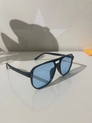 Svarta solglasögon med blå glas - Snygga solglasögon med svart båge och ljusblå glas. Modellen har en klassisk aviatorform med dubbla bryggor och tunna skalmar. Perfekta för dig som vill sticka ut med en cool och modern look.