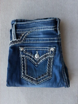Blåa miss me jeans  - Snygga blå jeans med markerade vita kontrastsömmar och dekorativa fickor bak. Jeansen har slitningar och tvättade detaljer för en cool look. Klassisk femficksmodell med raka ben och silverfärgade knappar. Perfekta för dig som gillar streetstyle. Har dock två små hål mellan benen som inte syns men går att enkelt fixa 