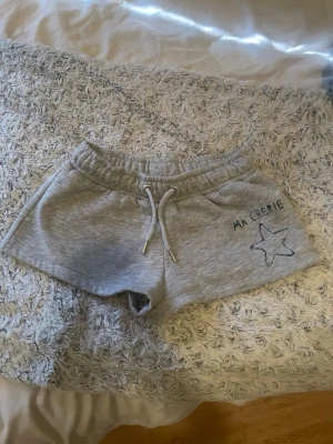 Grå mjukisshorts med snörning - Säljer ett par grå mjukisshorts med snörning i midjan och broderad text 'MA CHERIE' samt en stjärna på ena benet. Shortsen har en relaxed passform och är perfekta för chill dagar. Tillverkade i mjukt material för extra komfort.