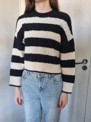 Svartvit randig stickad tröja kroppad  - Säljer en stickad tröja med breda svarta och vita ränder. Tröjan har rund hals, lång ärm och är något croppad i modellen. Materialet ser mjukt och fluffigt ut med ett diskret kabelstickat mönster. Perfekt för dig som gillar en avslappnad men trendig stil.