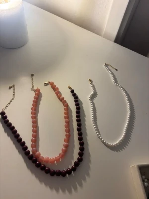 Pärlhalsband i olika färger - Säljer fyra snygga halsband med pärlor i olika färger: ett vitt, ett mörklila, ett ljusrosa och ett i guldton. Alla har rundade pärlor och justerbara kedjor i metall. Perfekta för att addera lite färg och stil till din outfit. Säljer var för sig eller tillsamman, skriv vilket smycke så ger jag pris ❣️