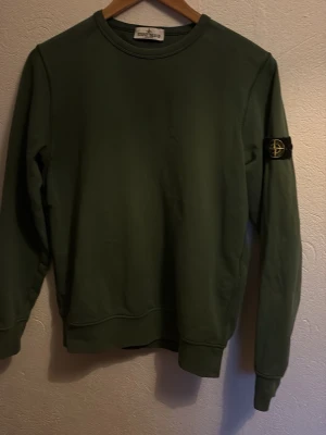 Grön sweatshirt från Stone Island Junior - Säljer en grön sweatshirt från Stone Island Junior med klassisk rund hals och lång ärm. Tröjan har den ikoniska Stone Island-loggan på vänster ärm och är tillverkad i mjuk bomull. Perfekt för en avslappnad och stilren look.