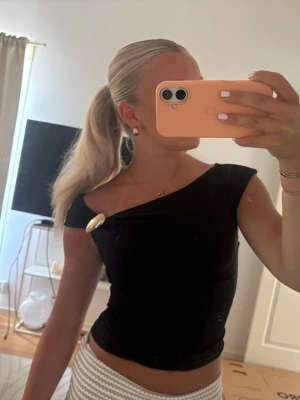 Svart topp med gulddetalj - Säljer en svart topp med en unik guldfärgad detalj på axeln. Toppen är croppad och har en tight passform som framhäver figuren. Perfekt för dig som vill ha något stilrent men ändå med en twist.