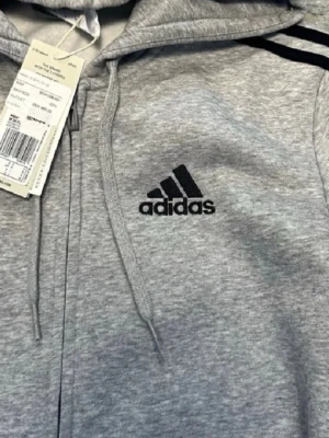 Grå adidas hoodie med dragkedja - Snygg grå hoodie från adidas med dragkedja framtill och klassiska svarta ränder längs ärmarna. Broderad adidas-logga på bröstet och justerbar huva med snören. Tillverkad i mjukt sweatshirtmaterial för en bekväm känsla.
