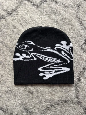 Svart mössa med grodmotiv - Säljer en svart mössa med ett stort vitt grodmotiv på framsidan. Mössan är mjuk och stretchig, perfekt för dig som vill sticka ut med en unik accessoar. Passar både till streetwear och chill vardagsstil.