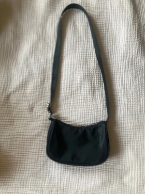 Svart axelremsväska i nylon - En stilren svart axelremsväska i nylon med enkel design och mjuk form. Väskan har en lång justerbar rem och stängs med dragkedja upptill. Perfekt för dig som gillar minimalistisk och tidlös stil.