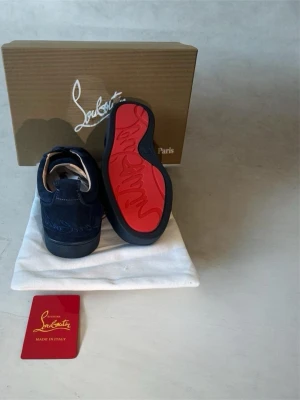 Mörkblå sneakers från Louboutin - Snygga mörkblå sneakers i mocka från Christian Louboutin med den ikoniska röda sulan och diskret logga på sidan. Skorna har klassisk låg profil, svarta snören och detaljer i svart gummi. Perfekta för dig som vill sticka ut med stil.