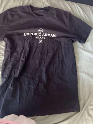 Svart Emporio Armani t-shirt - Snygg svart t-shirt från Emporio Armani med vit text och logga på bröstet. Klassisk rund halsringning och korta ärmar. Perfekt för dig som gillar stilrena och trendiga plagg med exklusiv känsla.