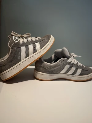 Grå Adidas Campus sneakers - Snygga grå Adidas Campus sneakers i mocka med klassiska vita ränder på sidorna och vit logga på hälen. Skorna har vita snören och en vit sula med gummidetalj. Perfekta för en clean och sportig stil. Kom gärna med prisförslag och frågor😀