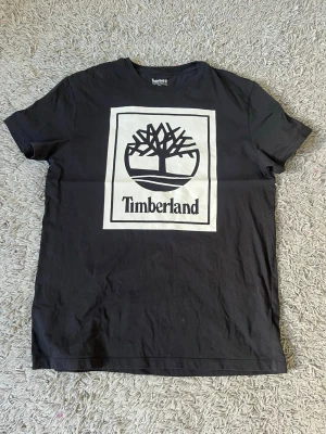Svart Timberland t-shirt med logga - Snygg svart t-shirt från Timberland med stor vit logga och trädmotiv på bröstet. Tillverkad i 100% ekologisk bomull för skön känsla och passform. Klassisk rund hals och korta ärmar, perfekt till jeans eller shorts.