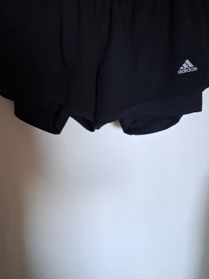 Svarta träningsshorts från Adidas - Säljer ett par svarta träningsshorts från Adidas med klassisk logga på ena benet. Shortsen har en sportig design med elastisk midja och innerbyxa för extra komfort. Perfekta för gymmet eller löprundan!
