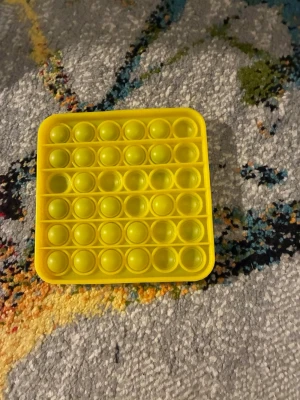 Gul fyrkantig pop it-leksak - En gul, fyrkantig pop it-leksak i mjukt silikonmaterial. Perfekt för att sysselsätta händerna och minska stress. Leksaken har flera rader med bubblor som kan tryckas in och ut för en tillfredsställande känsla.