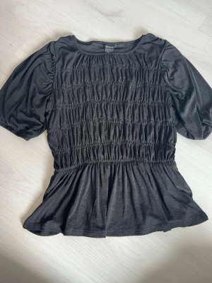 Svart puffärmad topp från Lindex - Snygg svart topp från Lindex med puffärmar och smockdetaljer över hela bröstet. Toppen har rund halsringning och en lätt utställd nederdel som ger en feminin siluett. Perfekt att styla med jeans eller kjol.