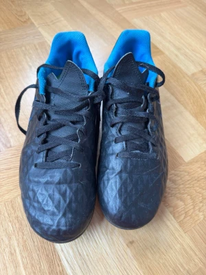 Nike Tiempo svarta fotbollsskor stl 38 - Säljer ett par svarta Nike Tiempo fotbollsskor med blå detaljer och innersula. Skorna har snörning, tålig ovandel i syntetmaterial med struktur för bättre bollkontroll och klassisk låg profil. Perfekta för dig som vill ha stilrena och funktionella skor på planen.
