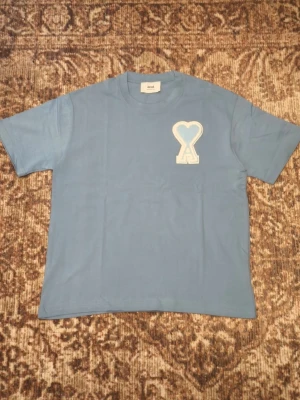 Blå t-shirt från Ami Paris - Snygg ljusblå t-shirt från Ami Paris med det ikoniska hjärtlogotypen broderad på bröstet. Klassisk passform och rund halsringning. Perfekt till jeans eller shorts för en clean och stilren look.