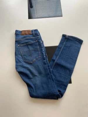 Blå jeans med raka ben, 30/34 - Säljer ett par klassiska blå jeans i modellen Evolve med raka ben och fem fickor. Jeansen har en snygg tvätt och bruna detaljer vid linningen. Tillverkade i bomull och perfekta för en avslappnad stil.