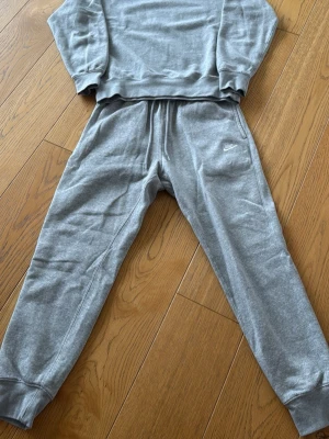 Grått loungewear-set från Nike - Mysigt grått loungewear-set från Nike med sweatshirt och matchande byxor. Setet har en enkel och stilren design med Nike-logga på byxan. Perfekt för chill dagar hemma. Tillverkat i mjukt material för extra komfort.